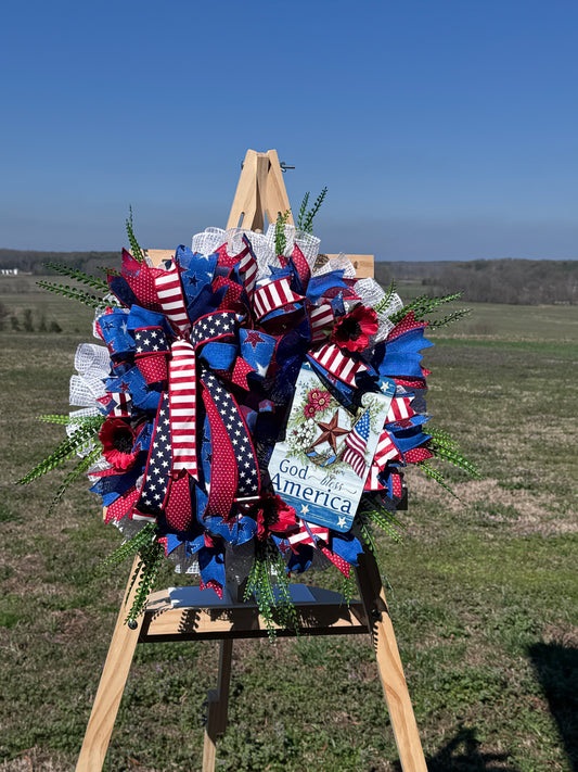 God Bless America wreath on a wooden easel outdoors, featuring patriotic red, white, blue ribbons and greenery.
