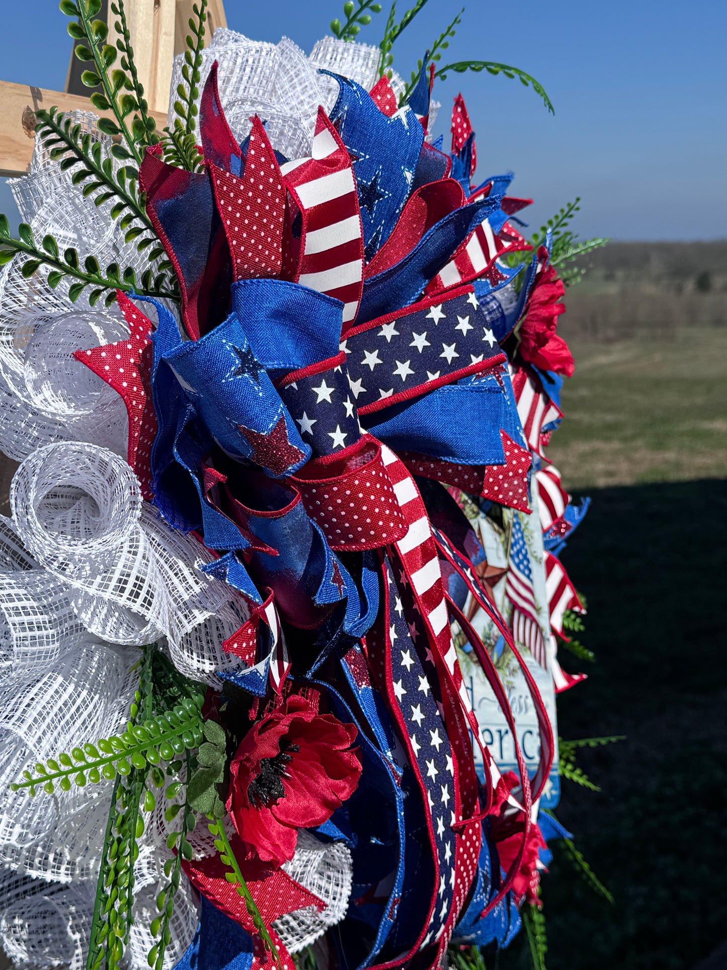 God Bless America patriotic wreath with stars-and-stripes ribbons, white mesh, and greenery.