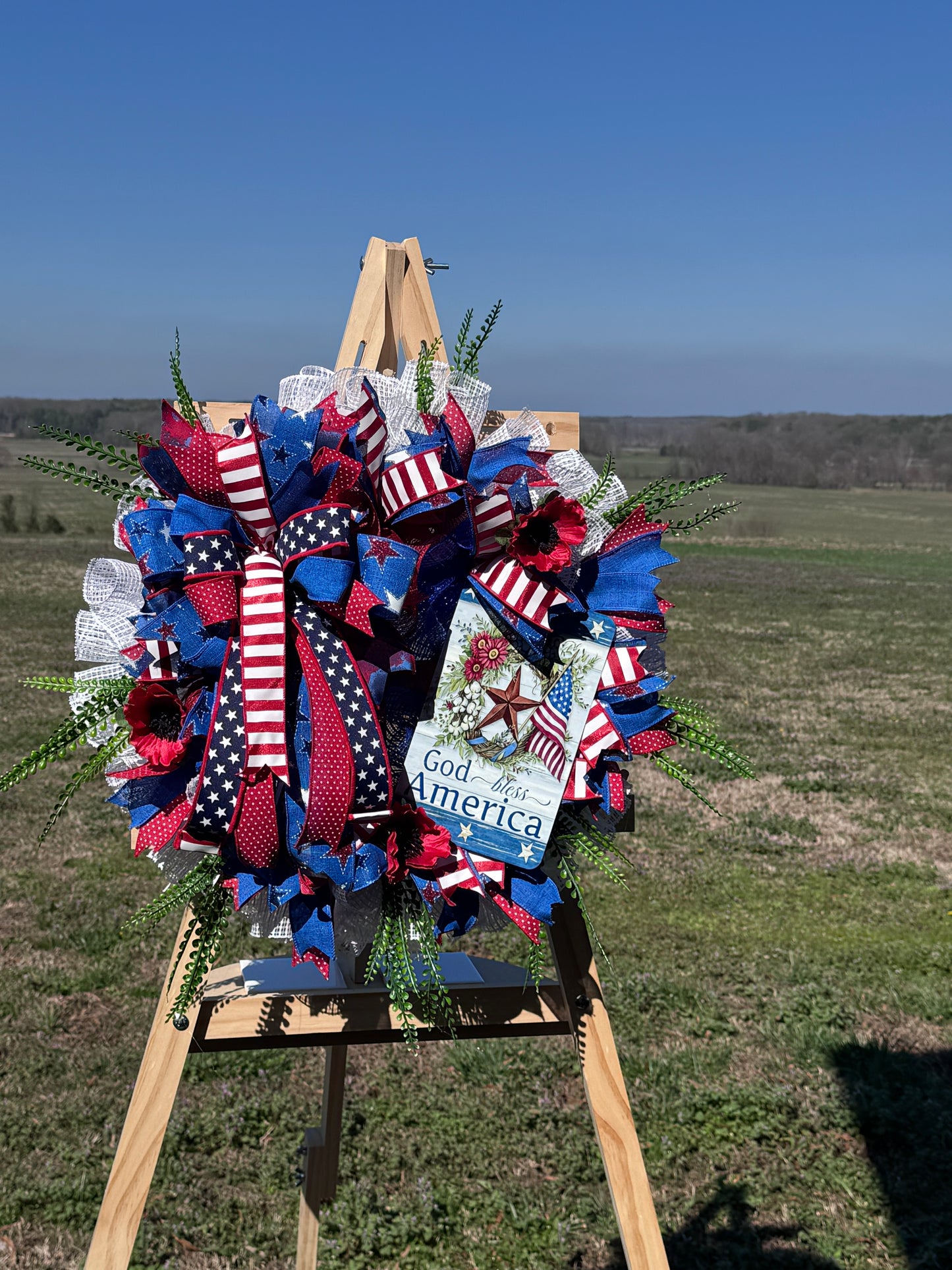 Patriotic wreath of red, white, blue ribbons with stars, greenery, and a 'God Bless America' tag on a wooden easel.