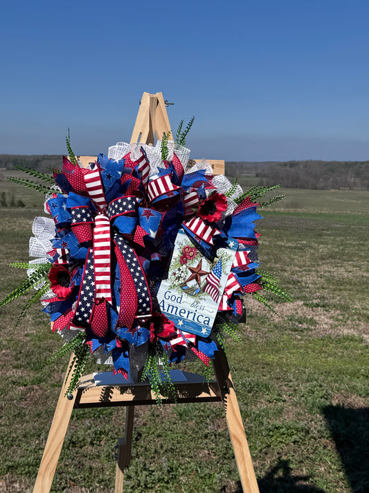 Patriotic wreath of red, white, blue ribbons with stars, greenery, and a 'God Bless America' tag on a wooden easel.